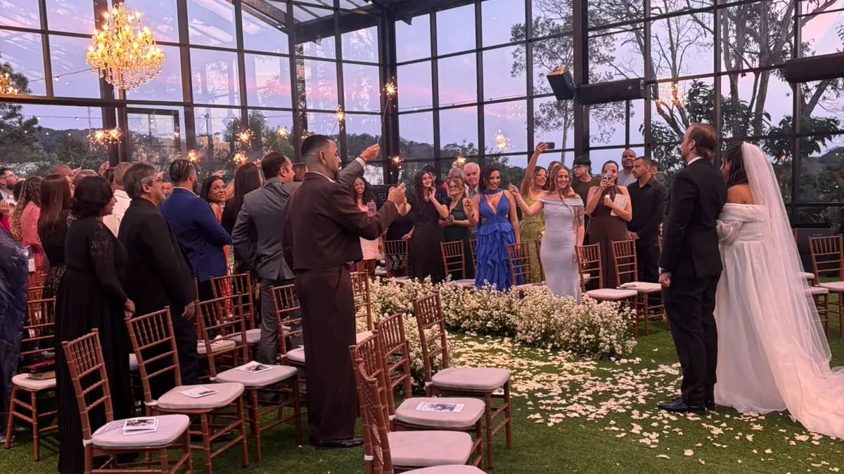 Wedding ceremony in a glass-walled hall with seated guests and the couple in a flower-lined aisle