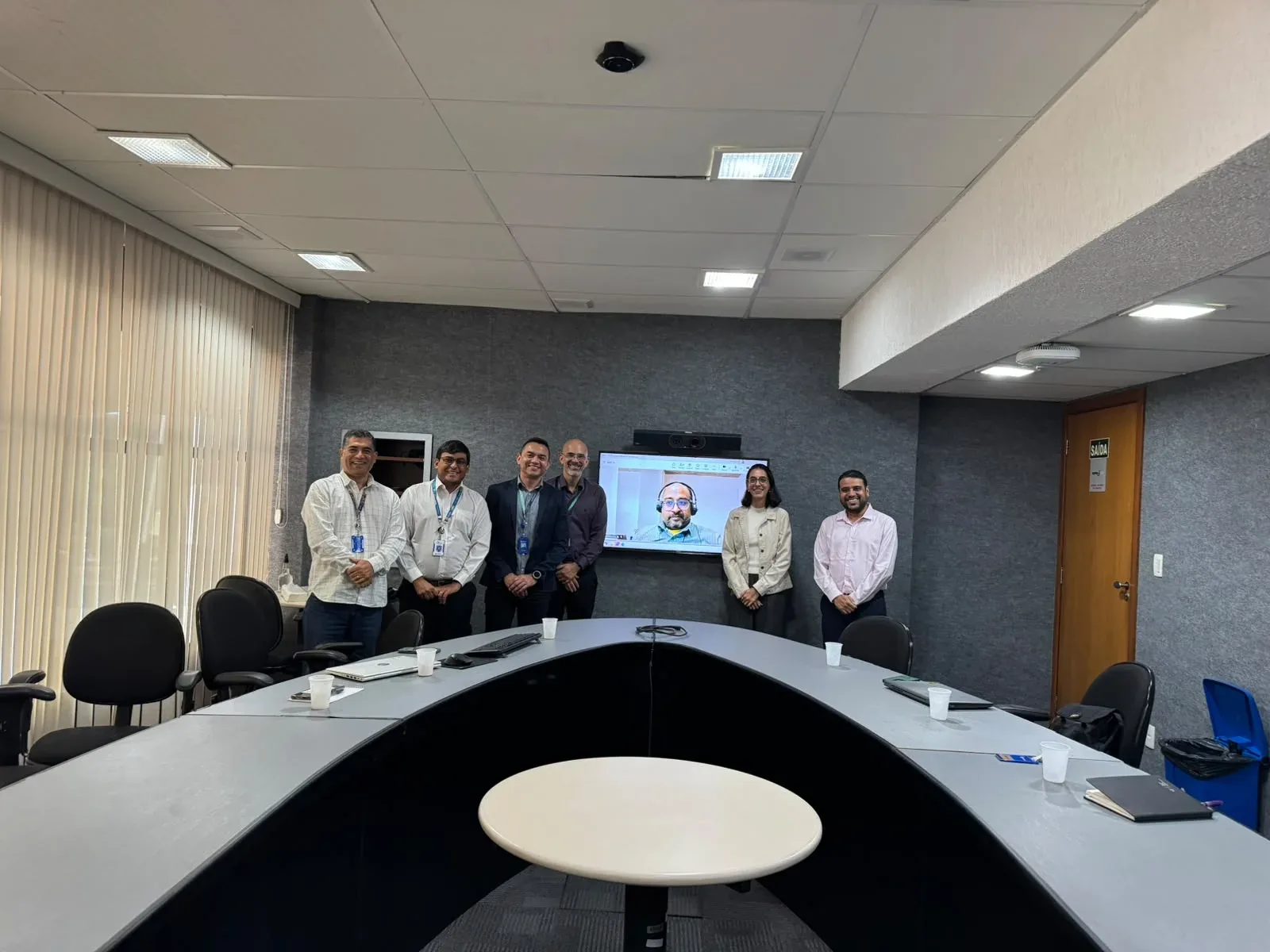 Meeting in Brasília with six participants standing and an Indian consultant joining by video call on screen